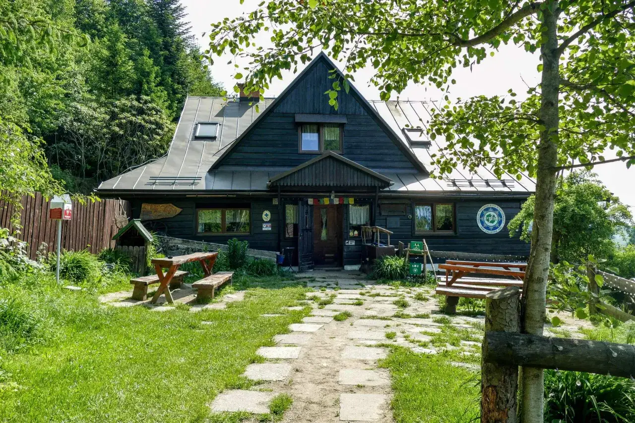 Urokliwa bacówka PTTK pod Bereśnikiem, otoczona zielenią, z drewnianymi ławami i stołami przed wejściem.