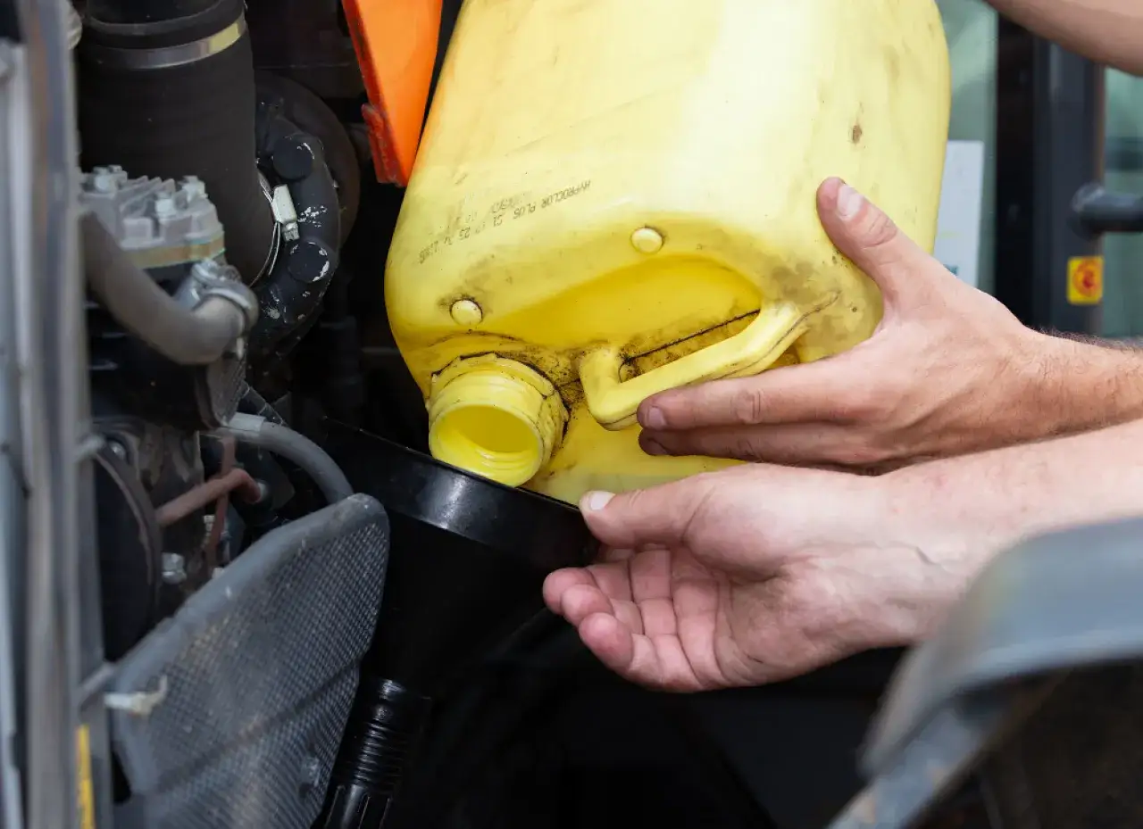 Dwie ręce przelewają żółty płyn z kanistra do czarnego lejka, obok silnika pojazdu.