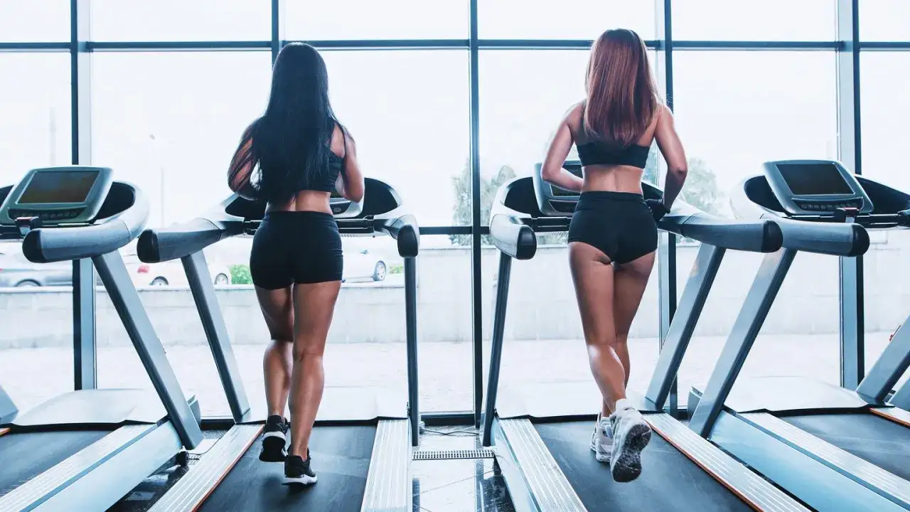Dwie kobiety biegnące na bieżniach w siłowni, widok od tyłu.