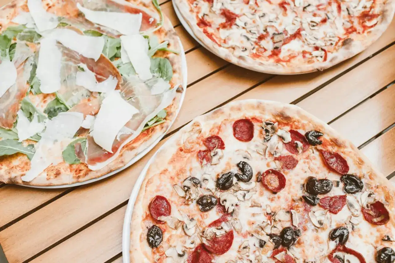 Trzy pizze na drewnianym stole. Jedna z szynką parmeńską i rukolą, druga z salami i pieczarkami, trzecia z pieczarkami. Szukasz alternatywy, jaki ser do pizzy zamiast mozzarelli?