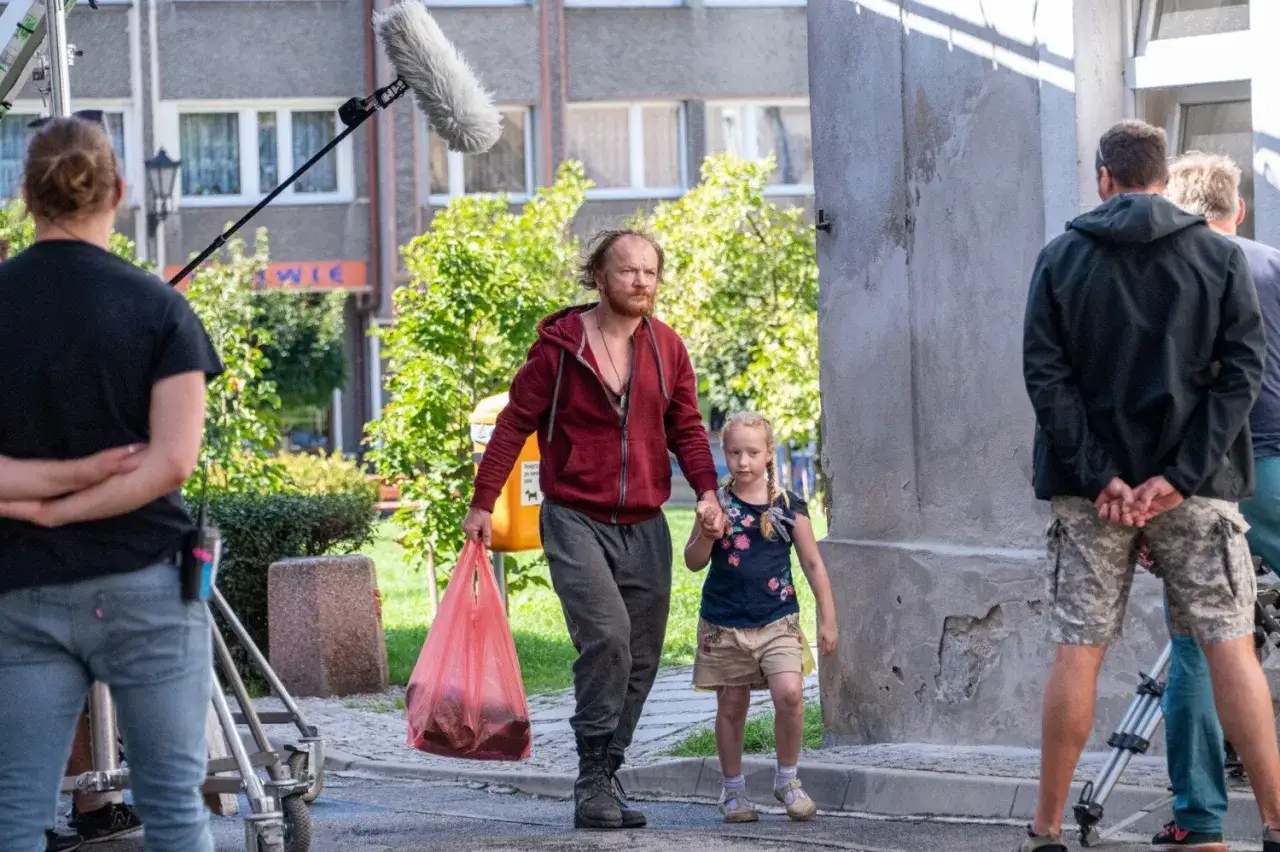 Mężczyzna z dziewczynką idą ulicą, za nimi widać ekipę filmową i mikrofon.