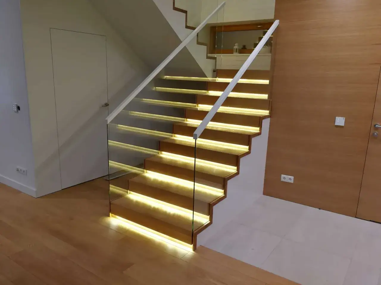 Nowoczesne schody dywanowe na beton z podświetleniem LED. Szklana balustrada i drewniane stopnie tworzą elegancką całość.