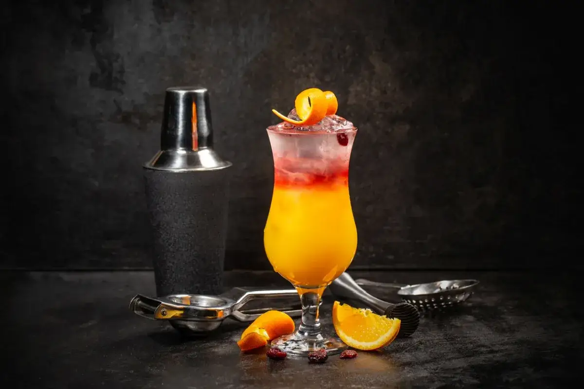Kolorowy drink Sex on the Beach z pomarańczą i żurawiną, obok shakera i akcesoriów barmańskich. Idealny przepis na lato!