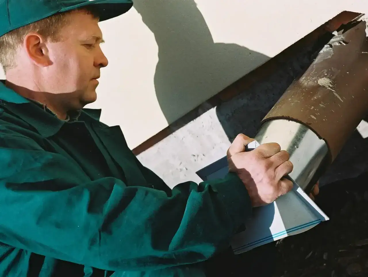 Mężczyzna w zielonej kurtce i czapce montuje rurę, która posłuży do doprowadzenia powietrza do kominka.