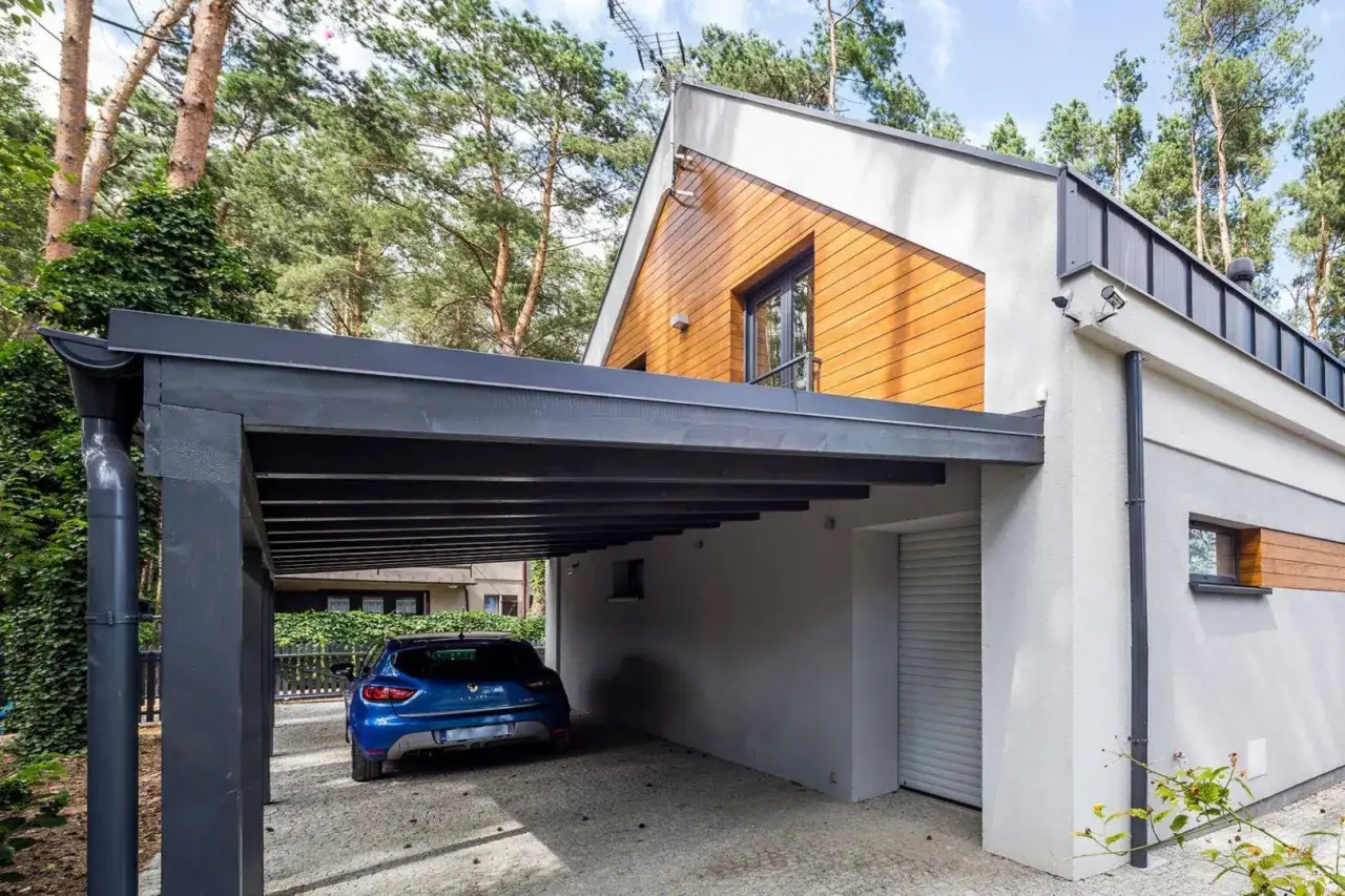 Nowoczesna wiata w granicy działki, zadaszająca niebieski samochód. Obok domu z drewnianą elewacją i garażem.