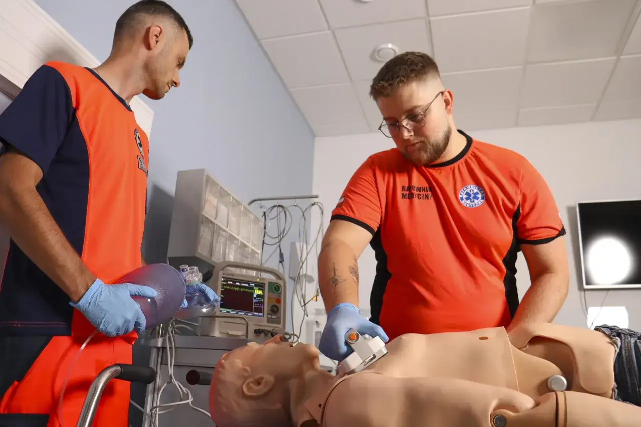 Dwóch ratowników medycznych ćwiczy resuscytację na fantomie, używając worka samorozprężalnego i defibrylatora.