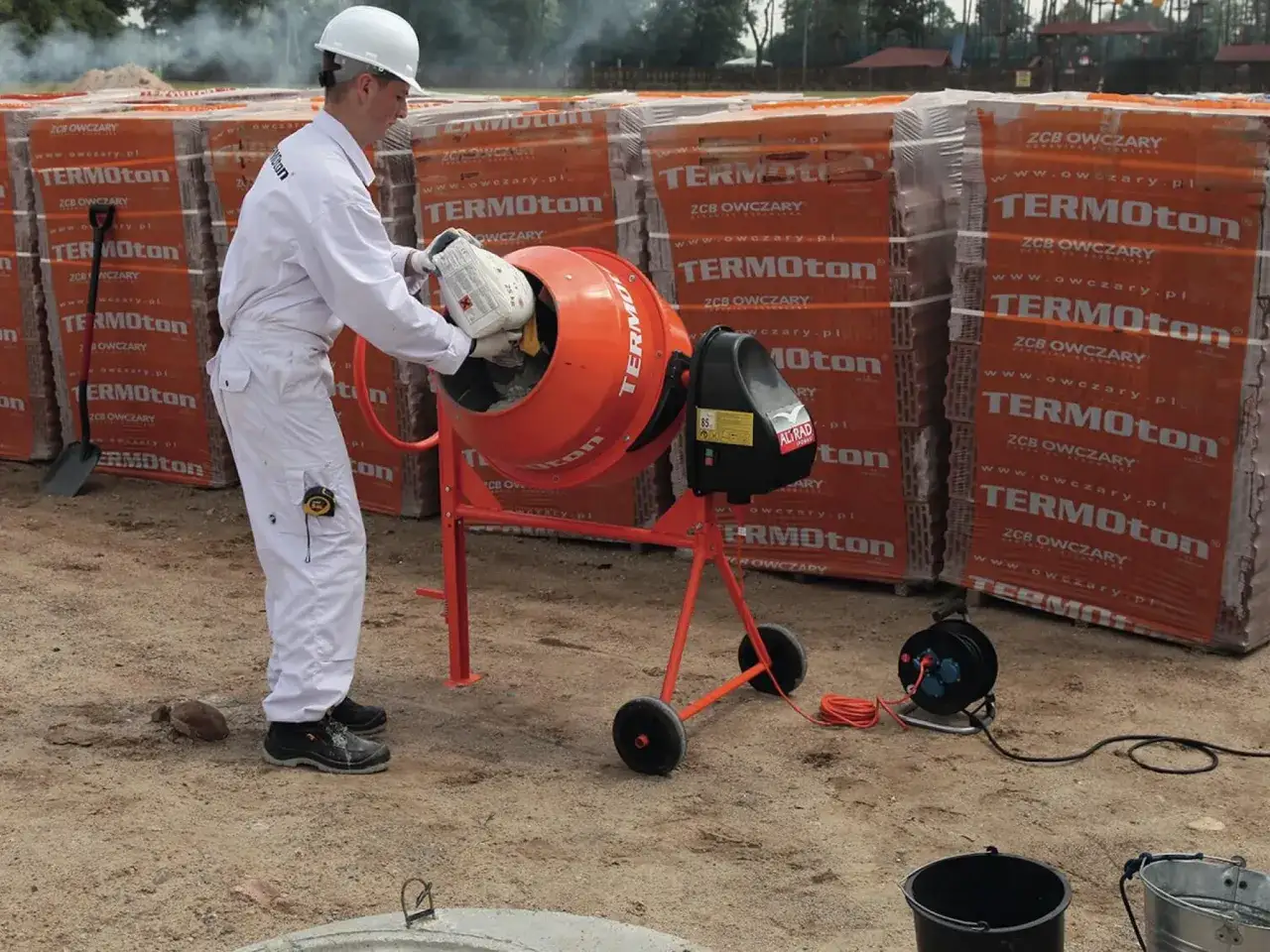 Mężczyzna w białym kombinezonie wsypuje składniki do betoniarki, przygotowując beton B30. W tle palety pustaków.