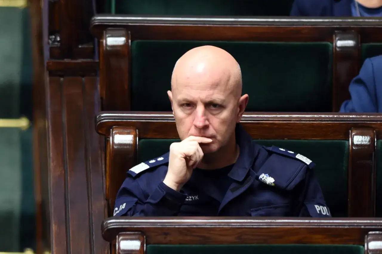 Łysy mężczyzna w mundurze policji siedzi w drewnianej ławie, zamyślony.