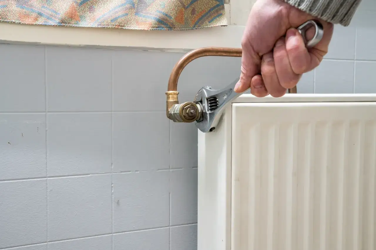 Dłoń dokręca kluczem nastawnym zawór przy grzejniku, pokazując jakie zawory do centralnego ogrzewania są potrzebne do montażu.