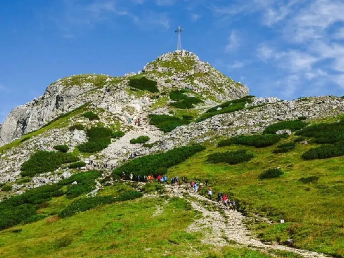 Grupa turystów wchodzi kamienistym szlakiem na zielone zbocze góry z krzyżem na szczycie.