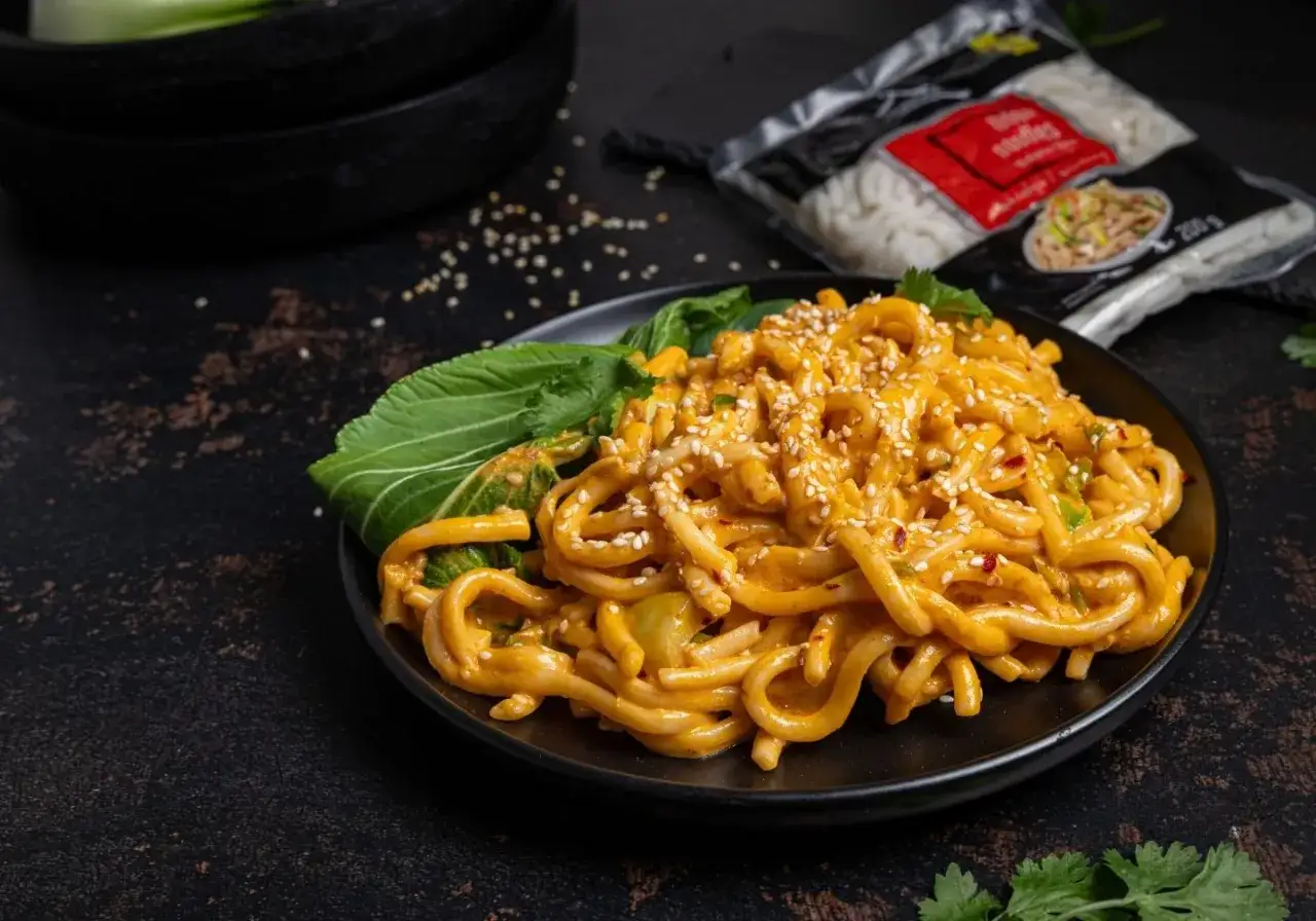 Grube, pomarańczowe kluski biang biang w sosie, posypane sezamem i chili, z zielonymi liśćmi bok choy.