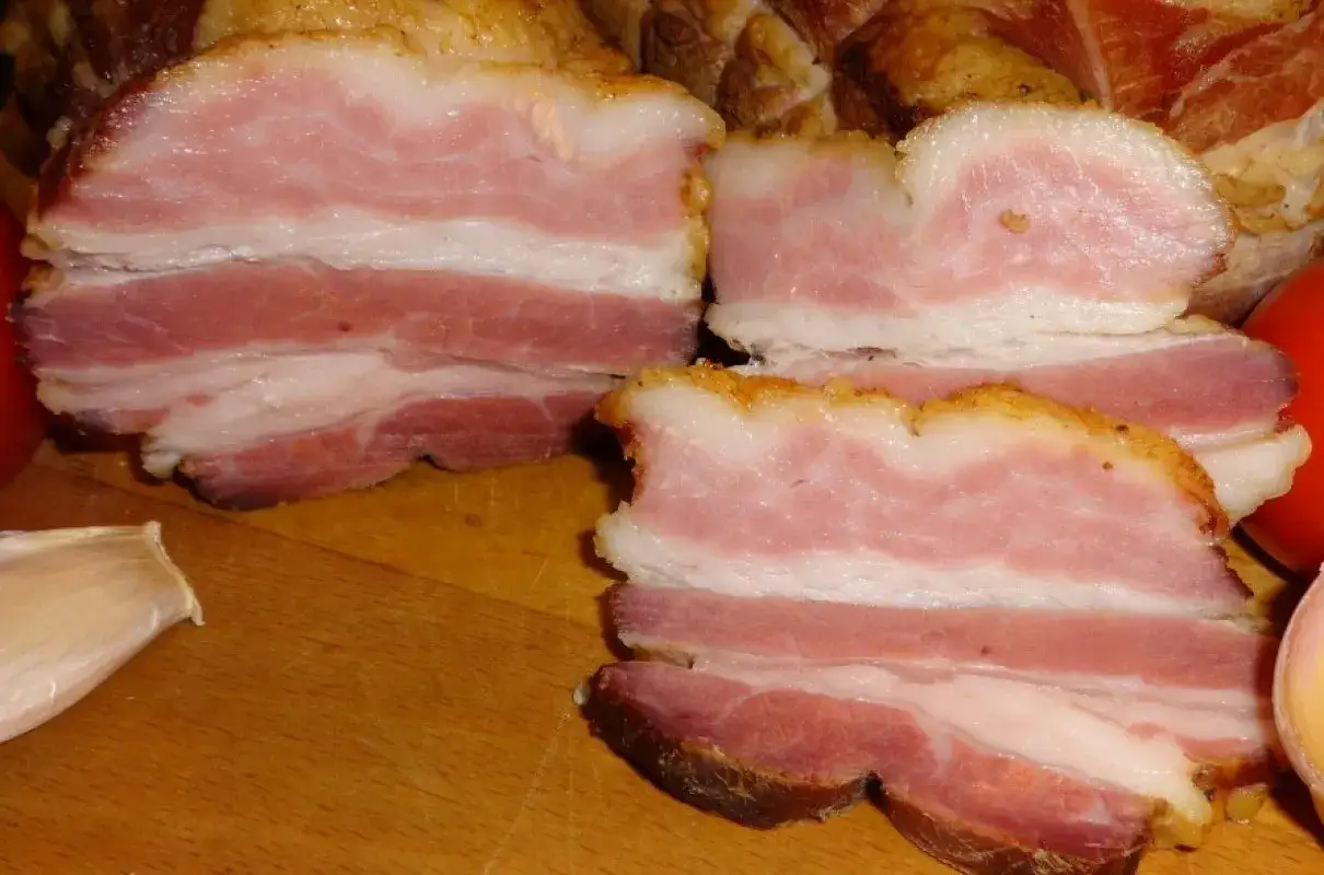 Kawałki wędzonego boczku na desce, obok czosnek i pomidor. Idealne do tego, jak sparzyć boczek wędzony.