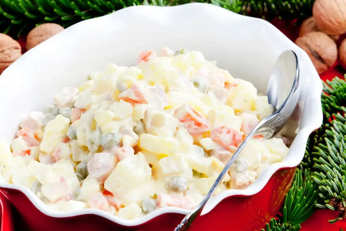 Sałatka jarzynowa w czerwonej misce z łyżką, ozdobiona gałązkami choinki i orzechami.