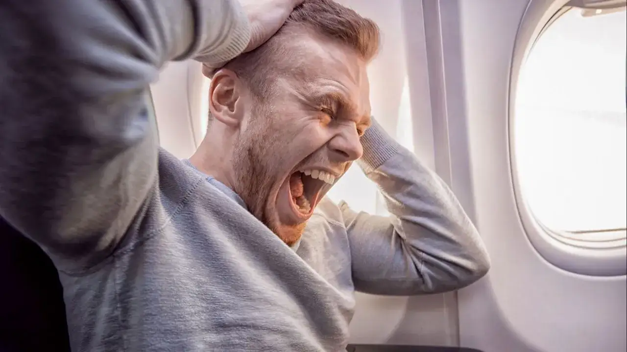 Mężczyzna w samolocie krzyczy z przerażenia, trzymając się za głowę.