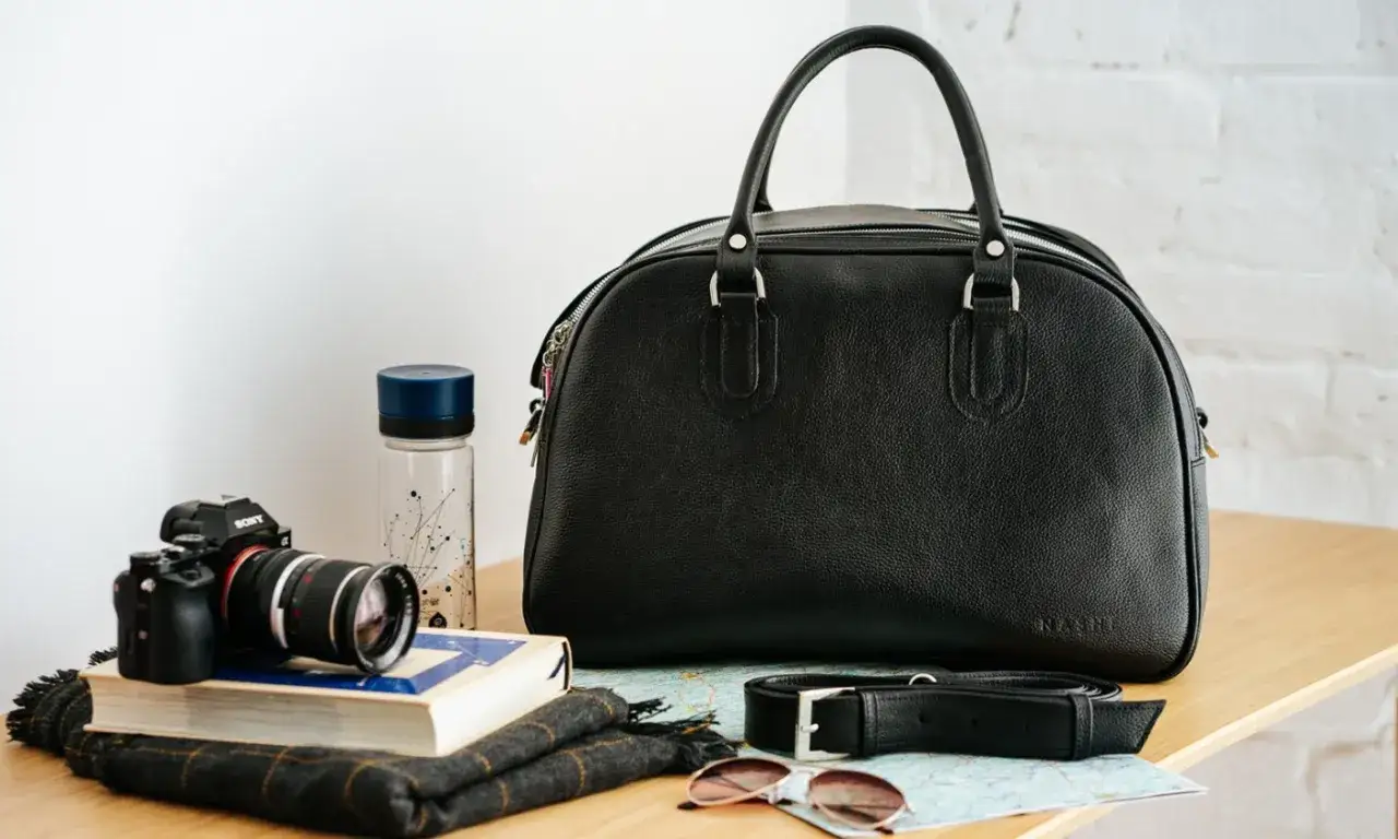 Czarna torba podróżna, aparat, książka, szalik, okulary przeciwsłoneczne i mapa.