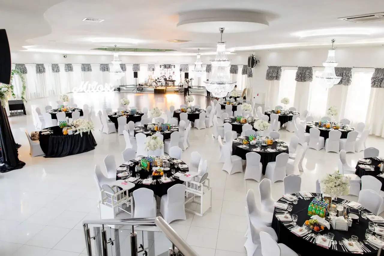 Elegancka sala weselna z czarnymi stołami, białymi krzesłami i kryształowymi żyrandolami.