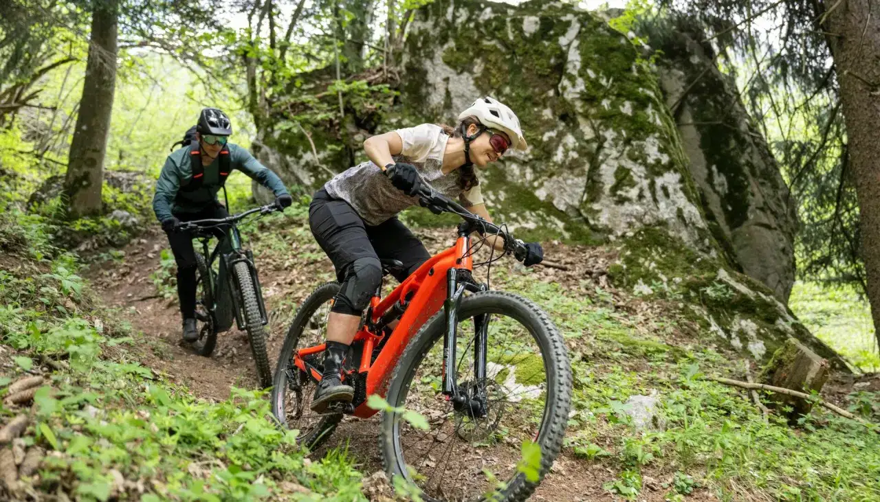 Dwie osoby na rowerach zjazdowych, jeden pomarańczowy rower bez amortyzatora z przodu, jadą po leśnym szlaku.
