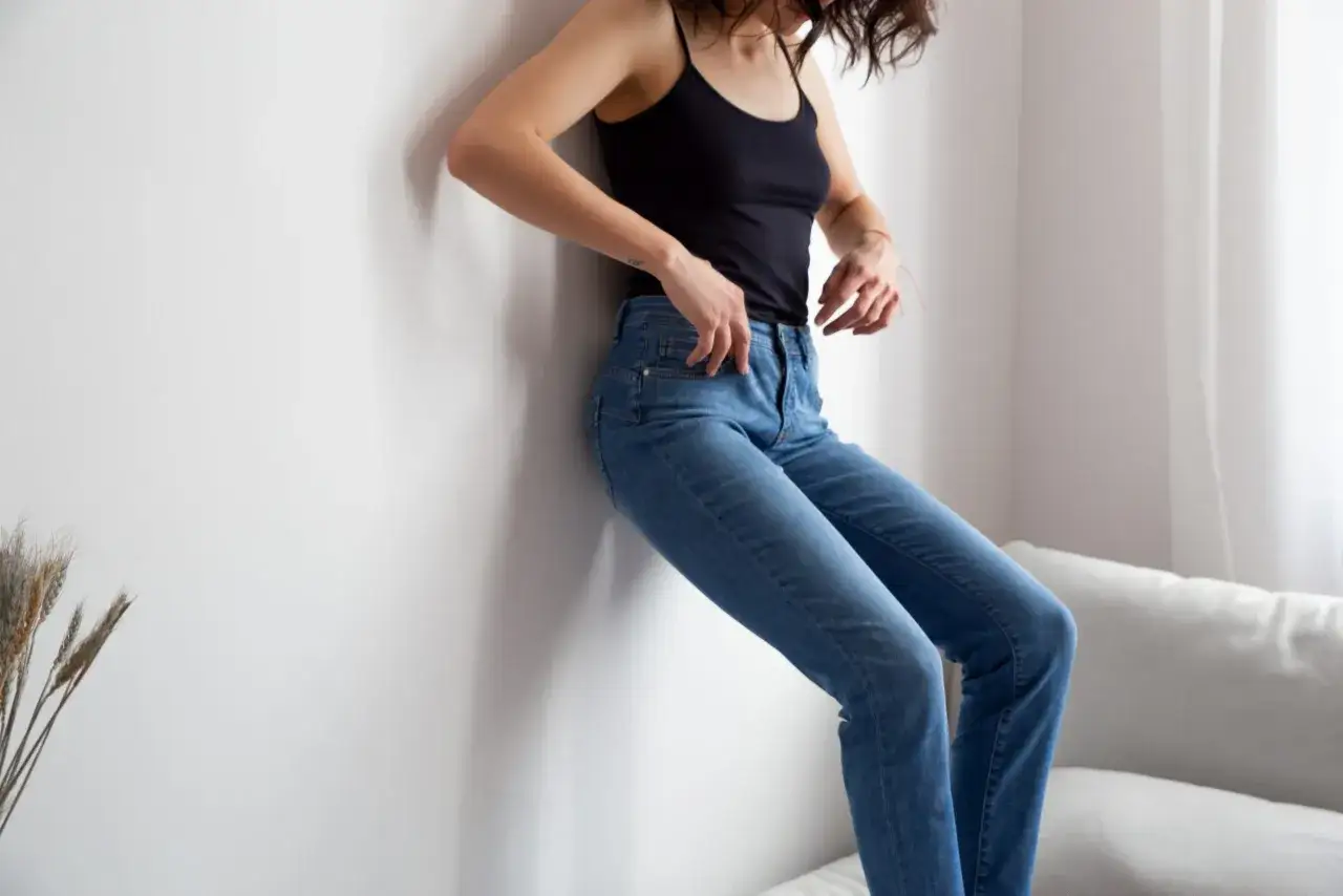 Kobieta w dżinsach opiera się o ścianę. Pokazano jej nogi, co może być pomocne, gdy zastanawiasz się, jak zmierzyć długość nogawki.