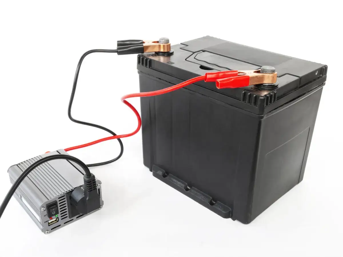Ładowanie akumulatora 100Ah: prostownik podłączony do czarnego akumulatora za pomocą czerwonych i czarnych klem.