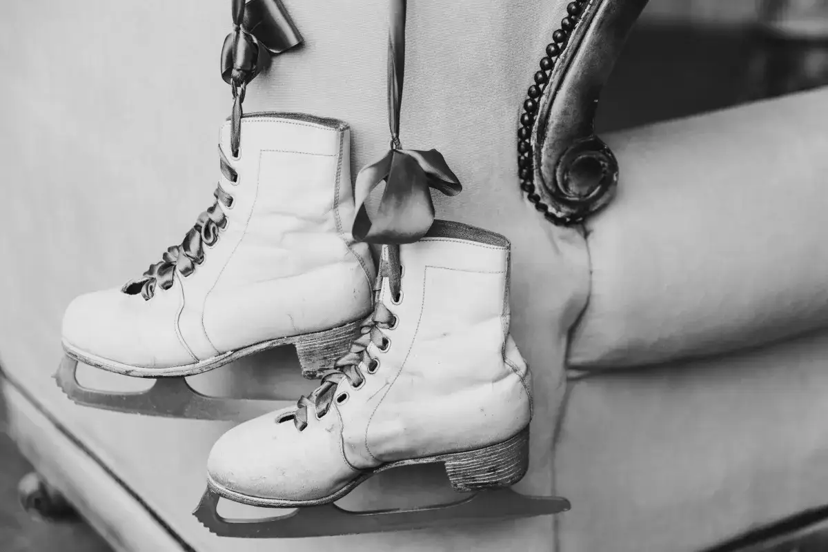 Białe łyżwy figurowe, ozdobione wstążkami, wiszą obok antycznego mebla. Przywołują wspomnienia o historii łyżwiarstwa figurowego.