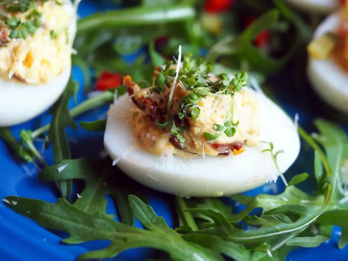 Jajka faszerowane suszonymi pomidorami, udekorowane kiełkami i rukolą, prezentują się apetycznie na niebieskim talerzu.