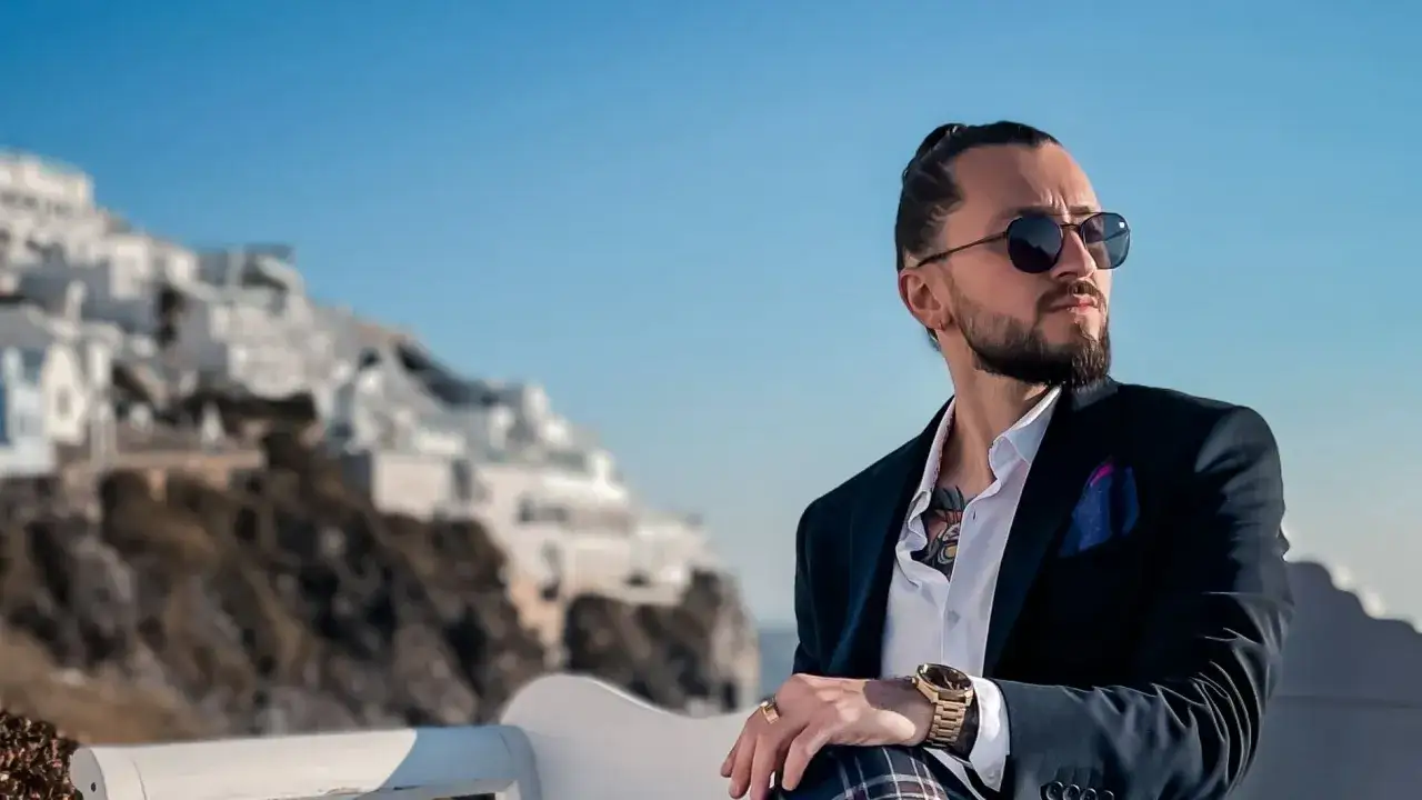 Elegancki mężczyzna w garniturze i z tatuażem na szyi, siedzący na tle białych domów. Czy prawnik może mieć tatuaż? Tak!