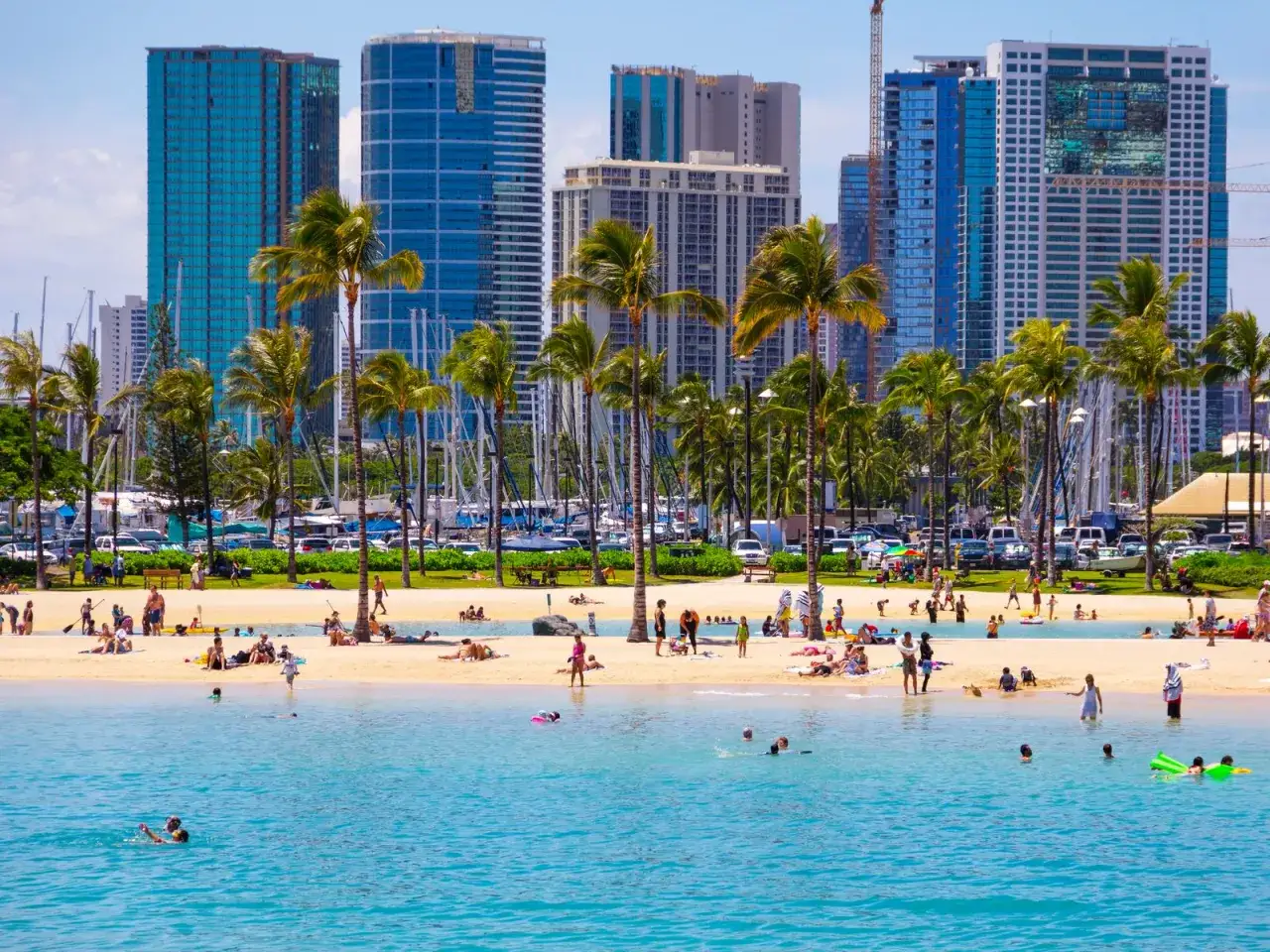 Plaża w Honolulu, palmy, turkusowa woda i wysokie budynki. Ludzie odpoczywają, pływają i opalają się. Zastanawiasz się, ile się leci do Honolulu?