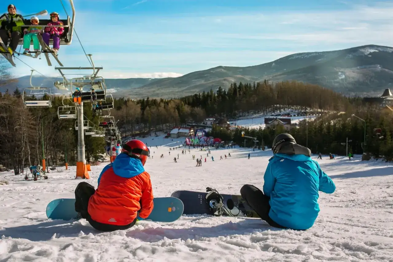 Dwaj snowboardziści odpoczywają na stoku, w tle widać wyciąg narciarski Karpacz i góry.