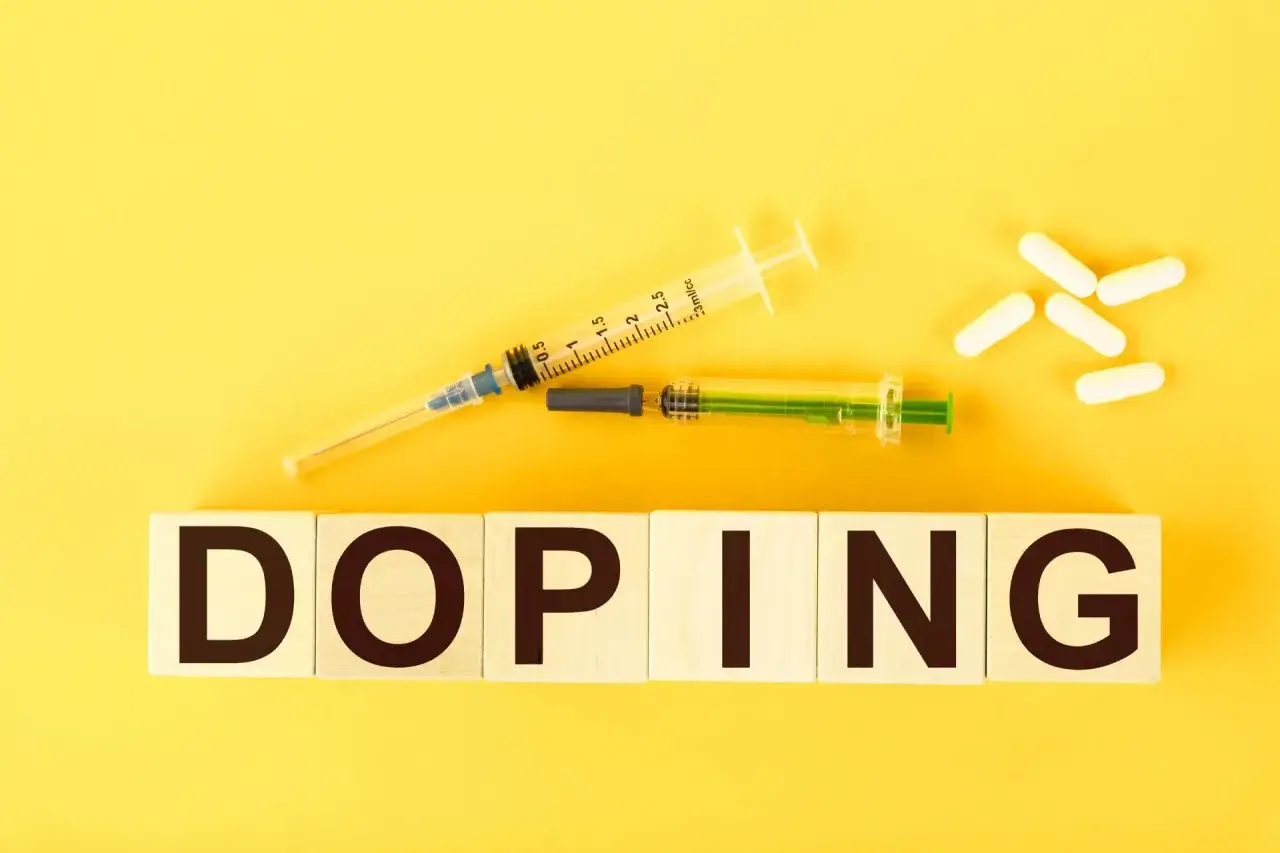 Dwa strzykawki i białe kapsułki na żółtym tle. Słowo "DOPING" napisane na drewnianych klockach.