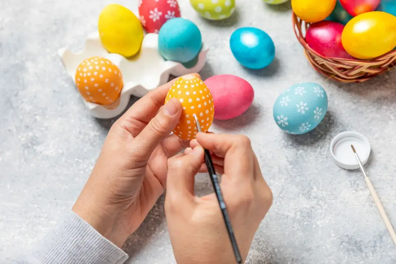 Ręce malują białe kropki na pomarańczowym jajku, tworząc piękne wzory na jajka wielkanocne. Wokół kolorowe pisanki.