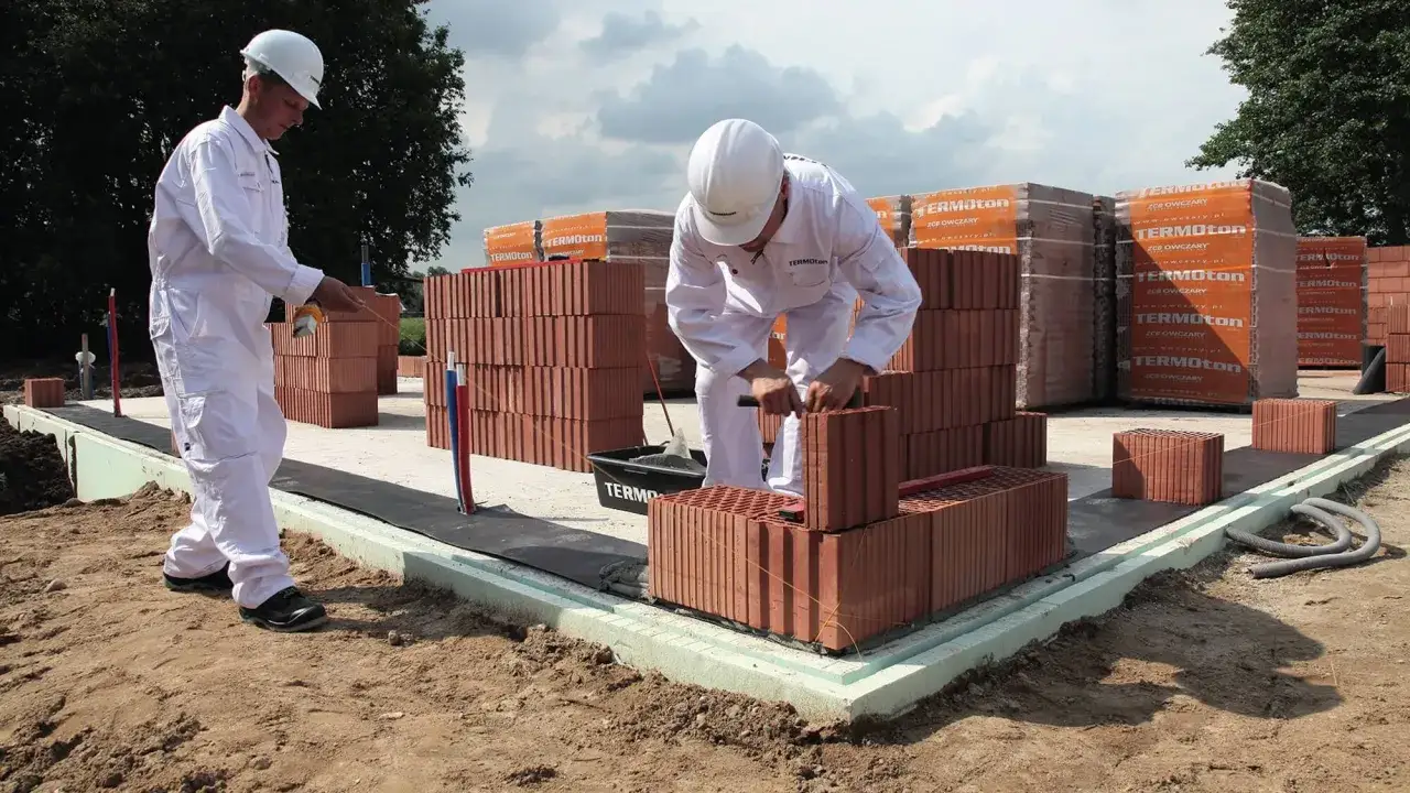 Dwóch budowlańców w białych kombinezonach układa cegły na fundamencie.