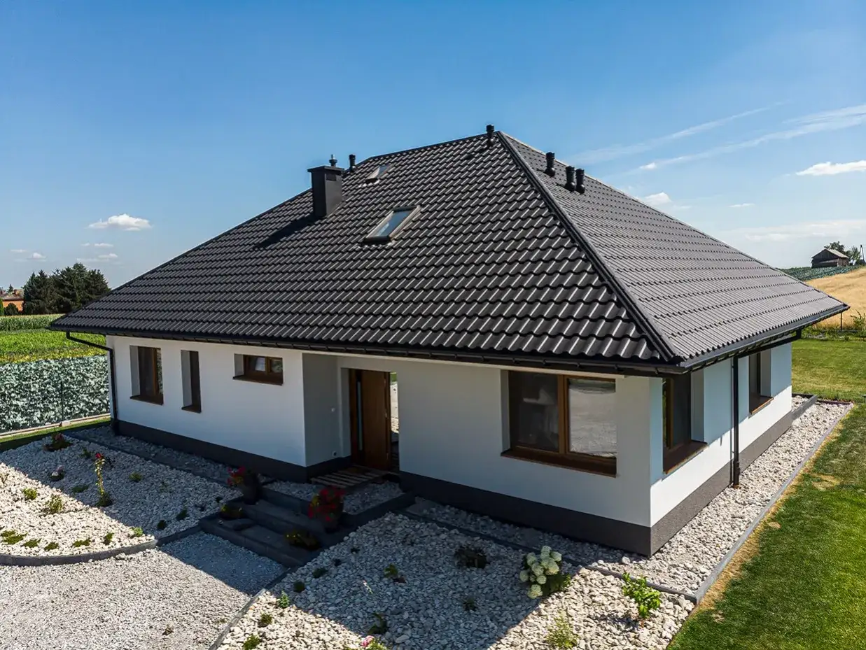 Nowy dom z czarnym dachem, gdzie kalenica jest najwyższym punktem dachu. Wokół zadbany ogród i pola.