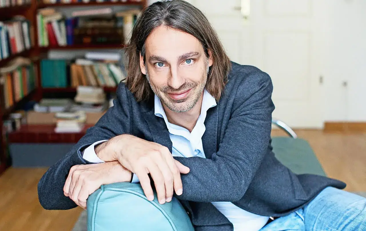 Richard David Precht sitzt lächelnd auf einem türkisfarbenen Sofa, im Hintergrund ein Bücherregal.