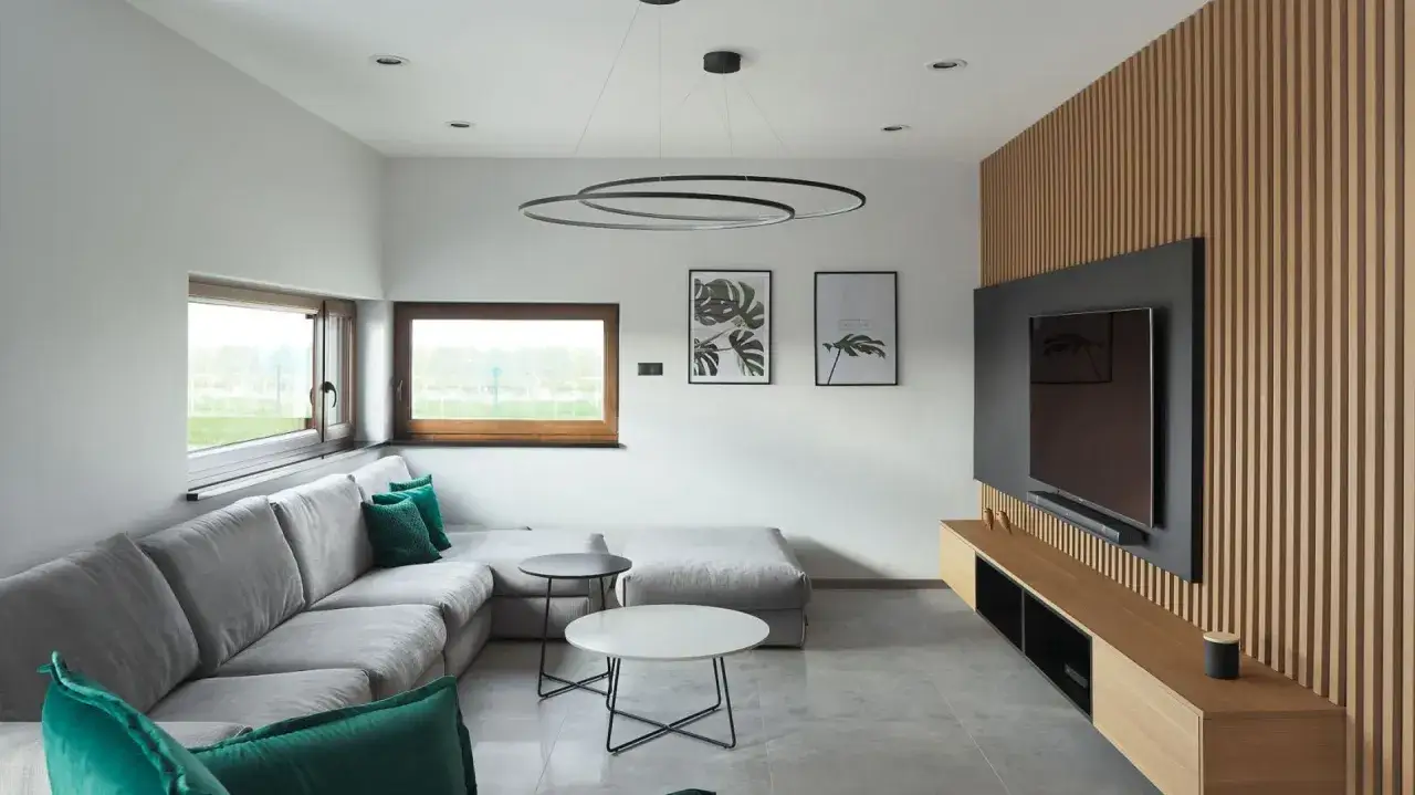 Nowoczesny salon z szarą kanapą i zielonymi poduszkami. Dekor na ścianie w salonie to drewniana boazeria i telewizor.