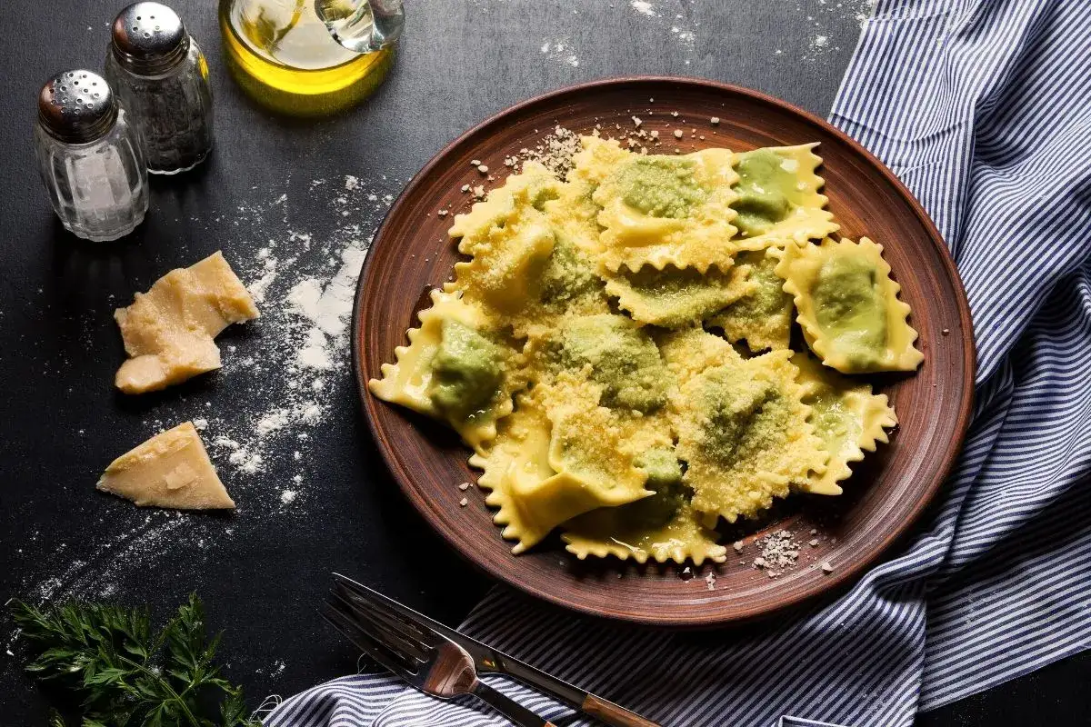 Pyszne ravioli z ricottą i szpinakiem, posypane parmezanem, na brązowym talerzu. Obok kawałki sera i przyprawy.