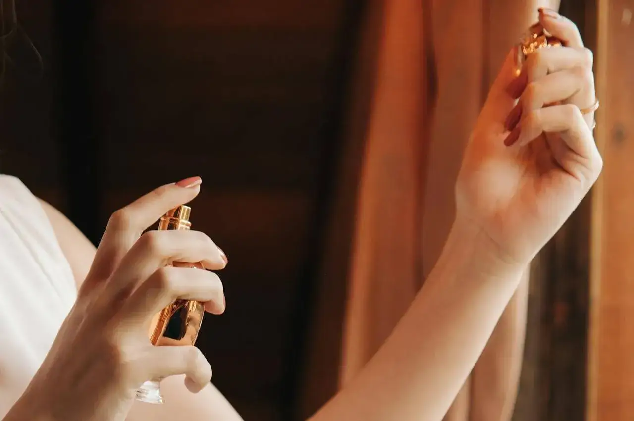 Dłoń kobiety trzymająca złoty flakonik perfum i aplikująca je na skórę.