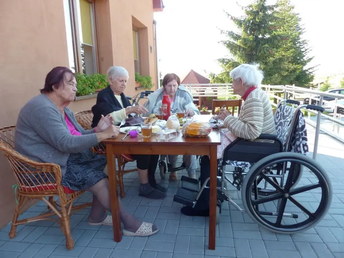 Cztery starsze osoby siedzą przy stole na zewnątrz, jedna na wózku inwalidzkim.