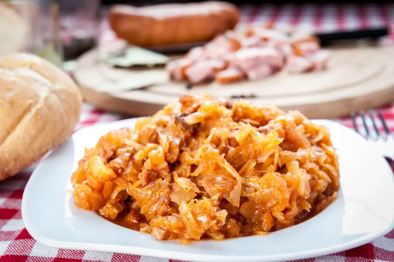 Pyszny bigos Ani, gotowany z pasją, na talerzu. Obok bułka i kawałki kiełbasy.