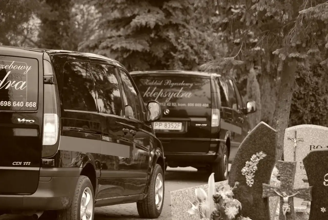 Dwa karawany pogrzebowe stojące obok siebie na cmentarzu, z widocznymi nagrobkami i drzewami.