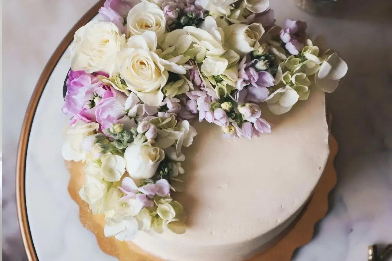 Elegancki tort z kremową polewą, ozdobiony bukietem żywych goździków, róż i hortensji w pastelowych kolorach.