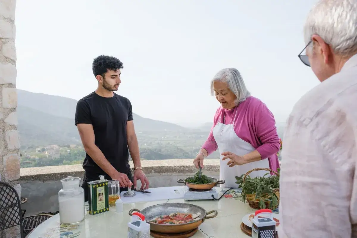 An older woman in an apron cooks greens while a young man and an older man stand nearby on a terrace with a scenic view.