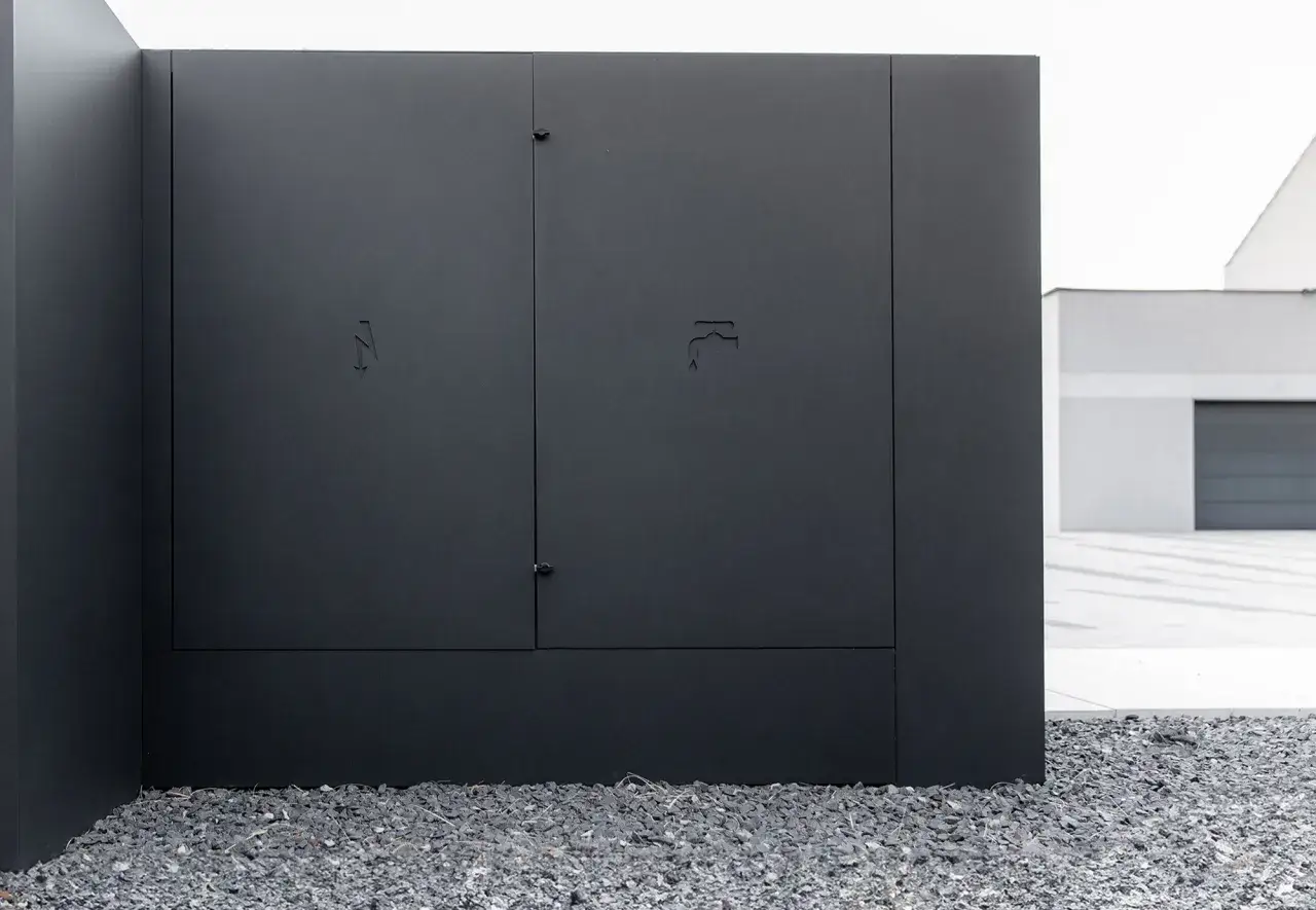 Nowoczesna, czarna skrzynka elektryczna w ogrodzeniu, z dyskretnymi symbolami błyskawicy i kranu.