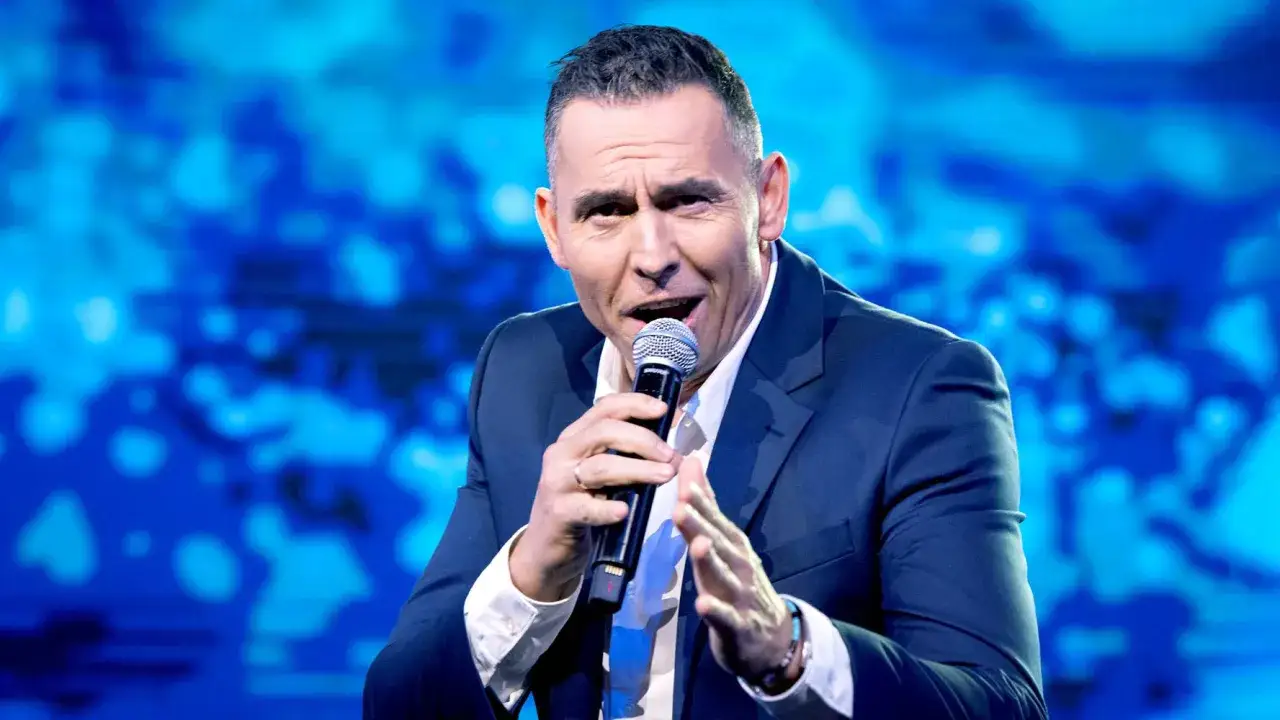 Mężczyzna w garniturze z mikrofonem, być może opowiada o tym, dlaczego Andrzej Kozłowski odszedł z kabaretu Pod Wyrwigroszem.