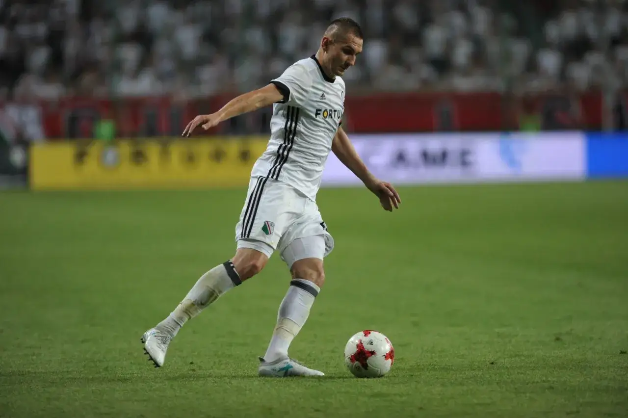 Piłkarz w białej koszulce z czarnymi pasami drybluje piłkę na zielonej murawie stadionu.