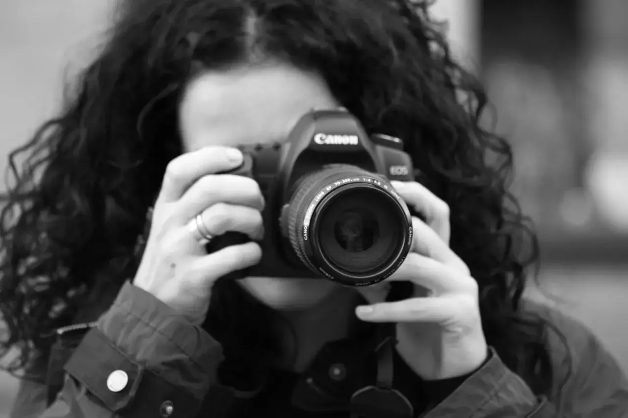 Czarno-białe zdjęcie kobiety z kręconymi włosami, która trzyma aparat Canon. To jej pierwsze szkolenie z fotografii.