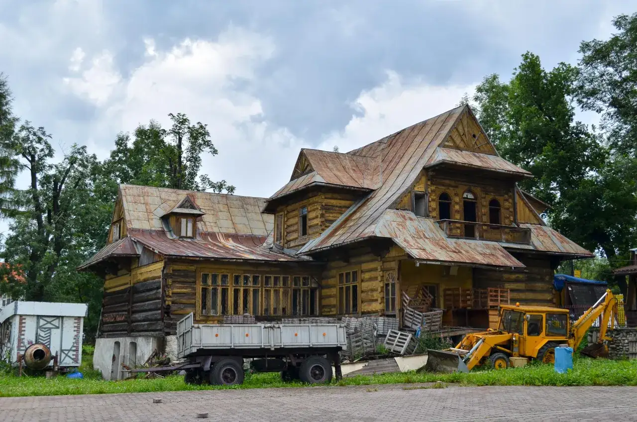 Drewniana chata w Bukowinie Tatrzańskiej, przypominająca starą turnię, z żółtą koparką i przyczepą na podwórzu.