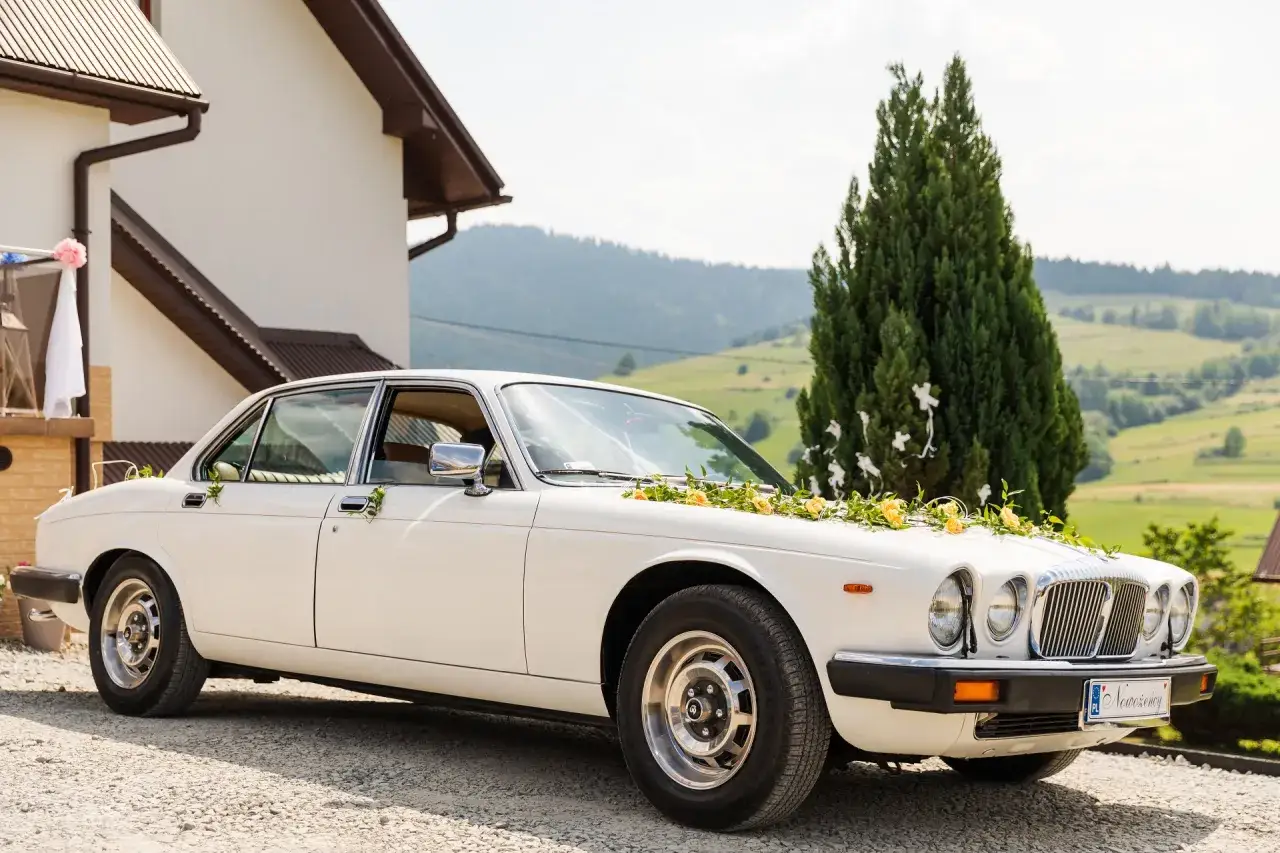 Biały samochód Jaguar ozdobiony kwiatami, zaparkowany przed domem, na tle górskiego krajobrazu.