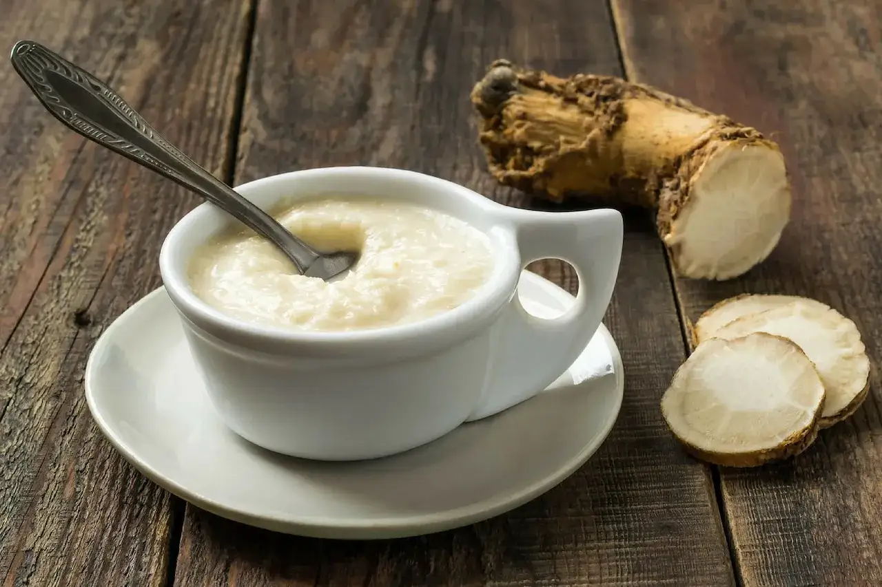 Biały sos chrzanowy w naczyniu, obok korzeń i plasterki chrzanu na drewnianym stole.