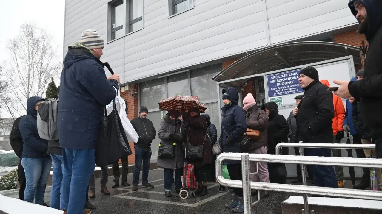 Grupa ludzi stoi przed budynkiem spółdzielni mieszkaniowej w deszczu, niektórzy pod parasolem.