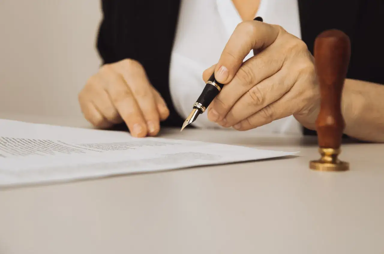 Dłoń trzymająca pióro nad dokumentem, obok pieczęć.